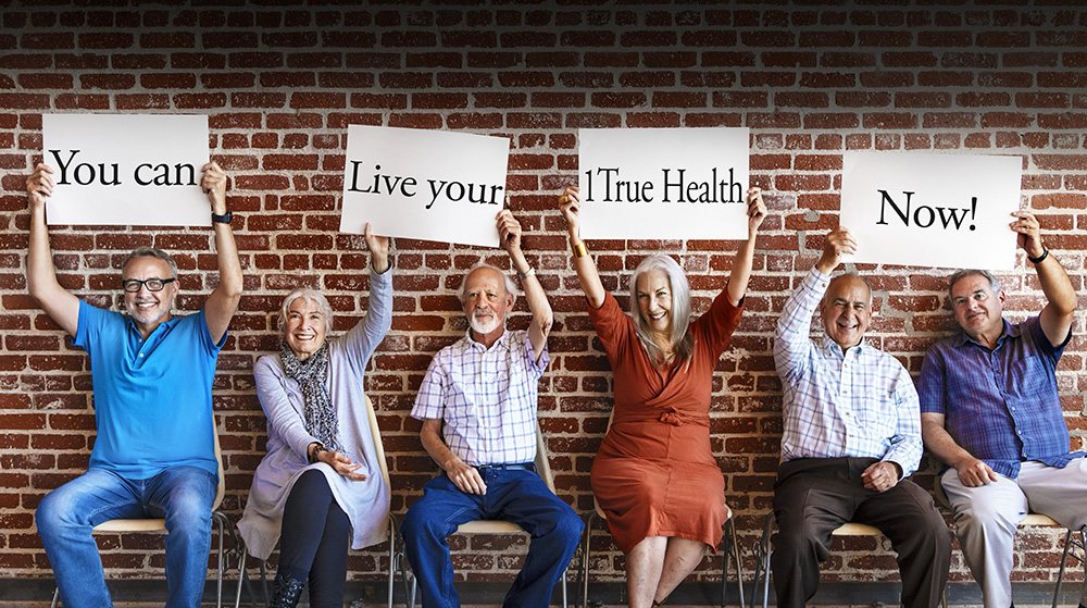 Six older adults seated against a brick wall, holding signs that read "You can — Live your — 1 True Health — Now!"